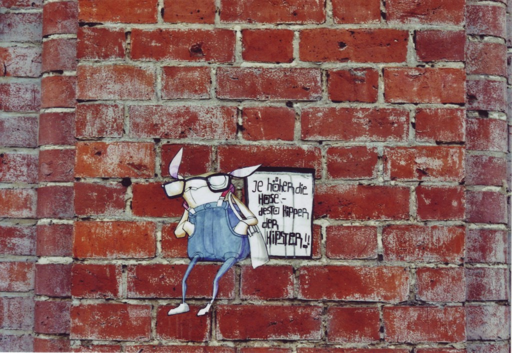 Street Art paste up auf einer Backsteinmauer: Eine Comicfigur, vielleicht ein Hase, mit Brille und Hose bis unters Kinn, Sprechblase: Je höher die Hose desto hipper der Hipster