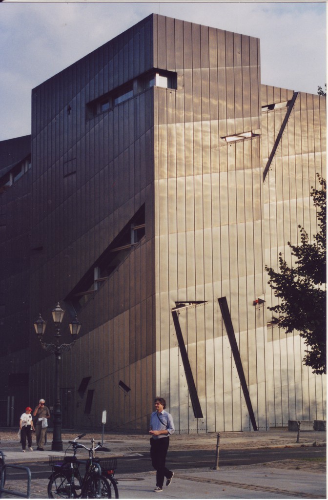 Fassade des von der Sonne angestrahlten Jüdischen Museums in Berlin, davor kreuzen einige Passanten die Strasse