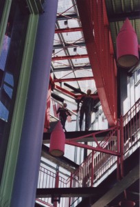 zwei Schaufensterpuppen stehen hoch oben in einem bunten, verwinkelten Glas-Stahl-Treppenaufgang
