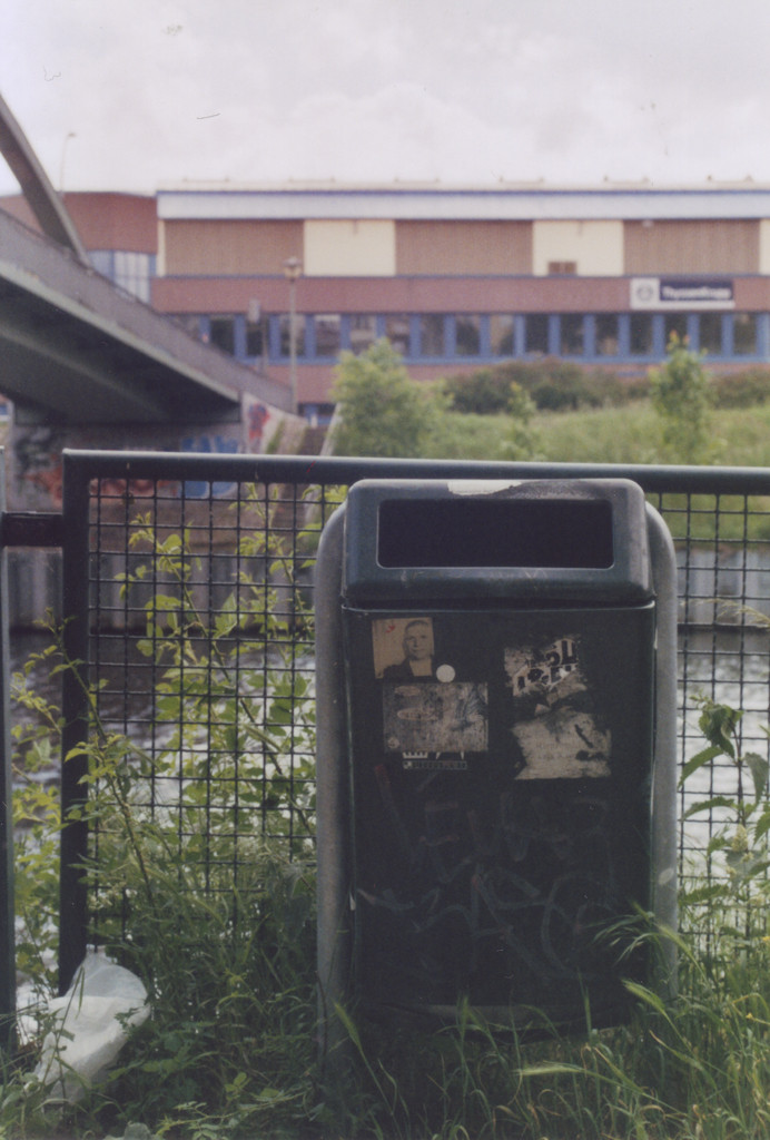 Ein Mülleimer mit mehren Aufklebern amn einem Kanal, im Hintergrund eine Brücke